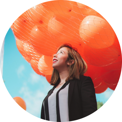 A round image of a smiling girl looking up towards a blue sky with orange balloons in the background