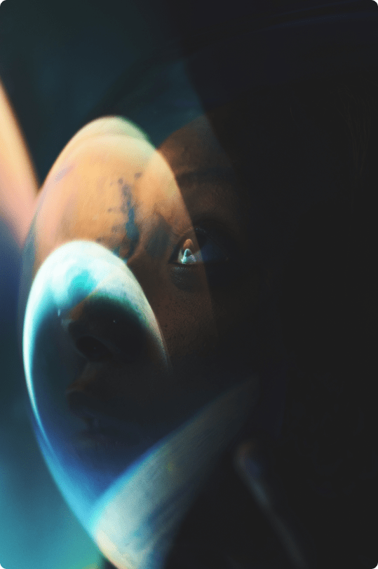 a woman in an astronaut helmet looking towards the earth with earth and space reflecting in the glass of her helmet.